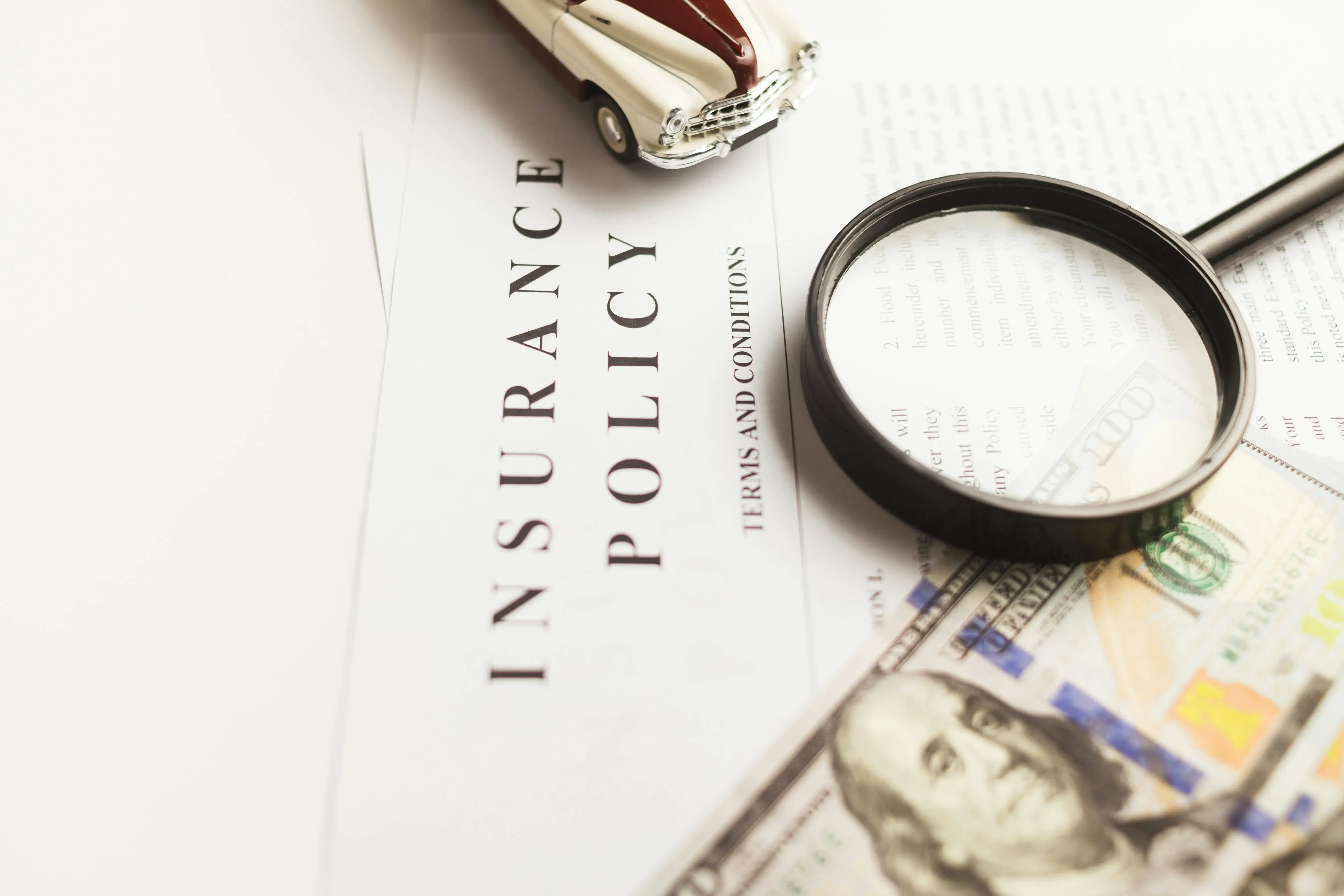 an insurance document on a desk. There is a toy car, a magnifying glass, and a hundred dollar bill stacked on top. What is the insurance policy hiding?
