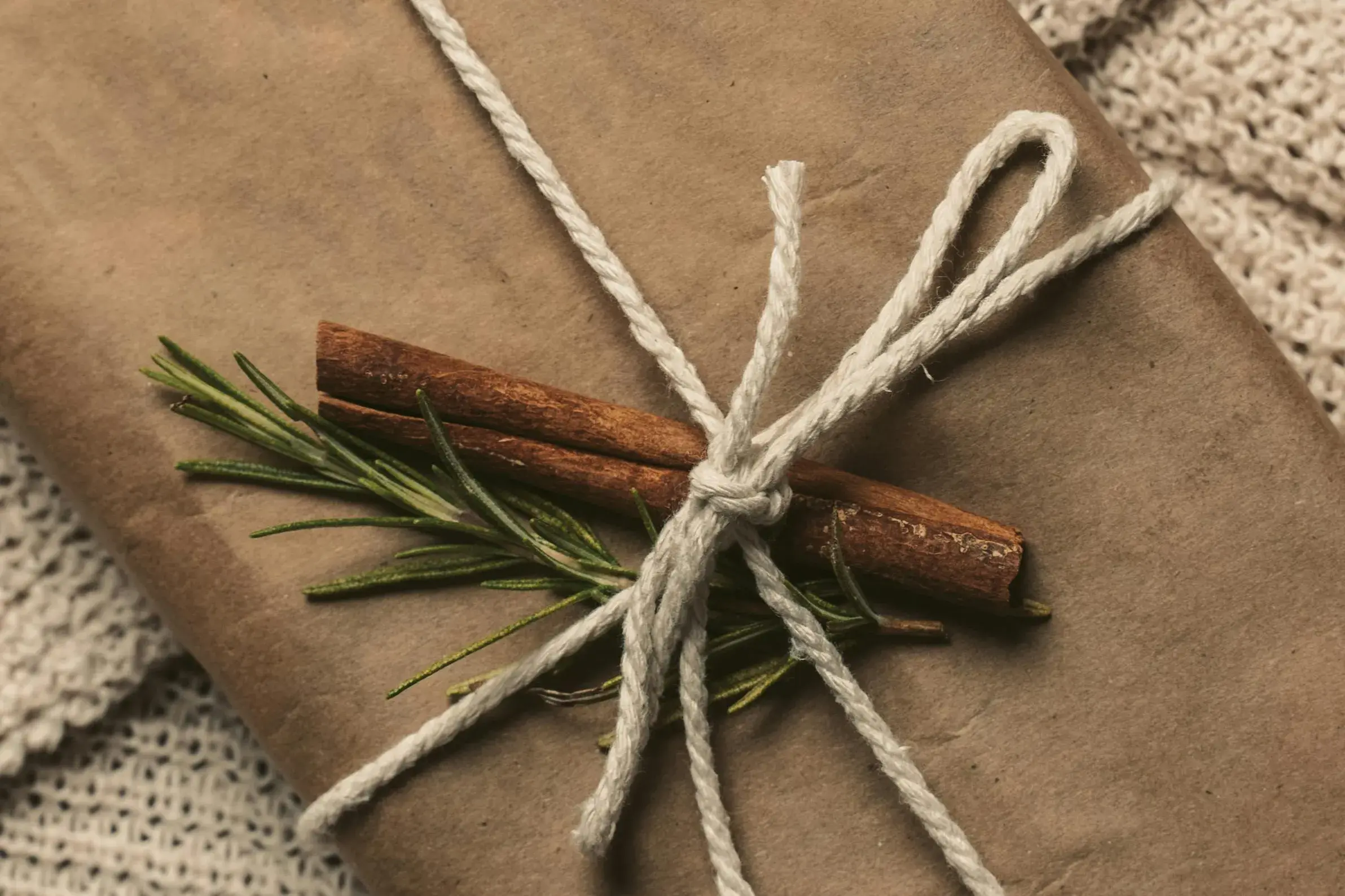 "Gift wrapped in brown kraft paper tied with natural cotton twine and decorated with cinnamon sticks and fresh rosemary sprigs on a knitted blanket.