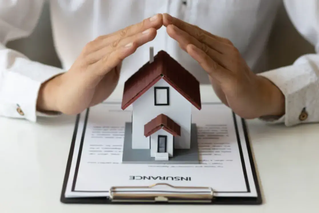 A model home sits on a clipboard. Hands are shielding the top, like an additional roof.
