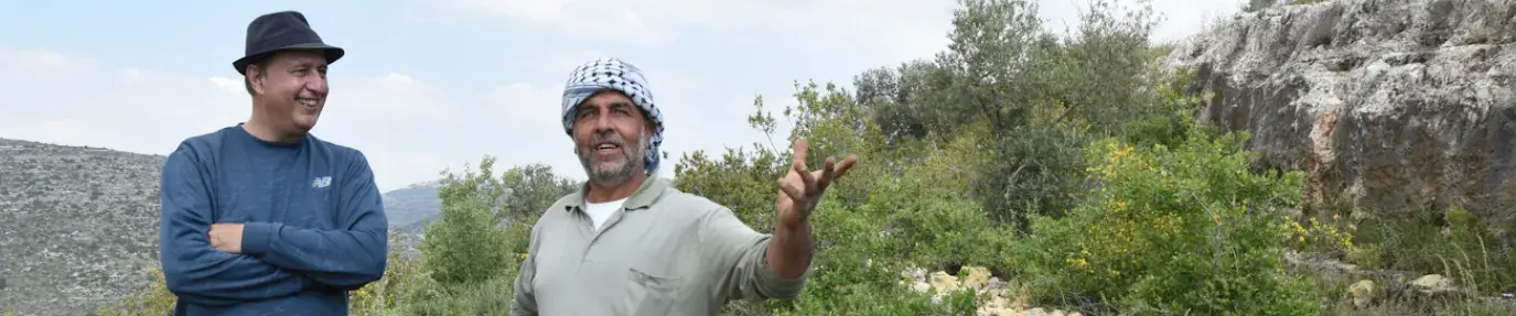 Nasser Abufarha stands to the left on a grassy hill in Palestine, arms crossed in a long-sleeve blue shirt and black hate. On the right is a local Palestinian farmer, in a grey shirt and turban, as they discuss regenerative agriculture.