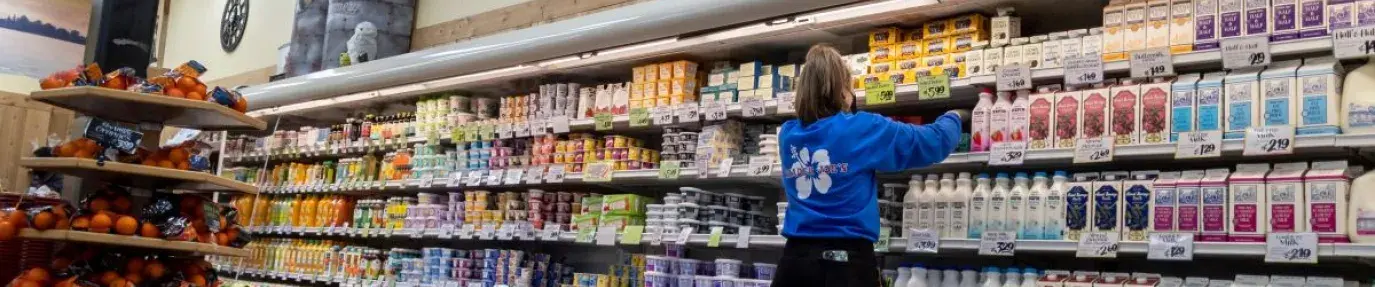 A woman in a blue trader joe's employee shirt organizing the shelves in the dairy section.
