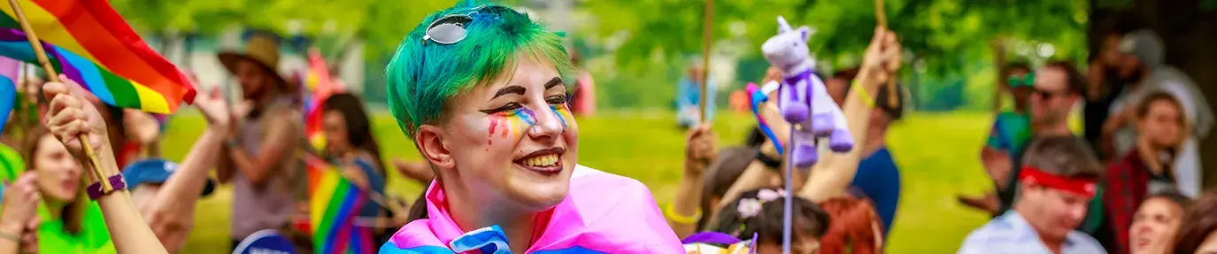 Happy person with rainbow makeup wearing a trans flag as a cape in a parade of rainbow flags.