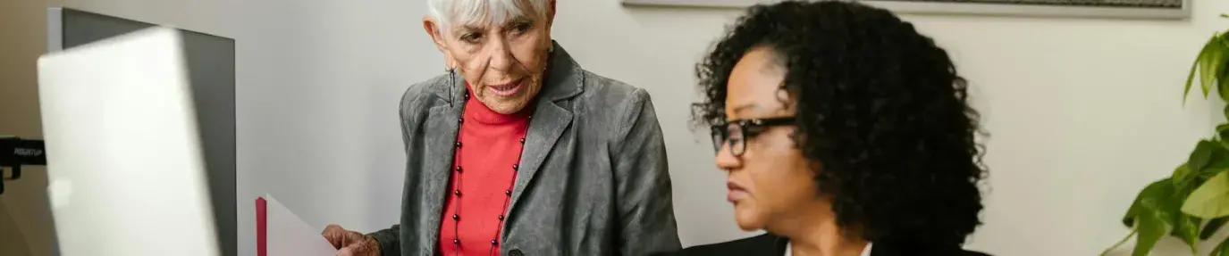 A Black woman sits at a desk in front of a computer desktop and keyboard with a clipboard in her hands. An older white woman in a grey blazer stands beside her with a folder of papers. Tax Planning Sustainable Growth Business.
