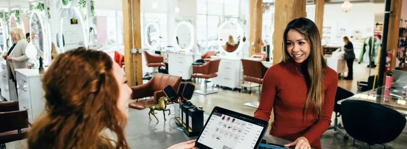 red-haired salon worker with a big smile standing behind counter accepting payment from a woman customer in a red sweater with a big smile, businesses that do a sustainability audit are on their way