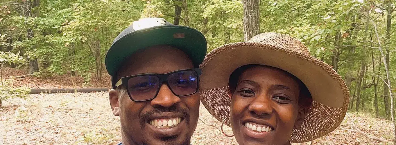 Osei Doyle and Brendalyn King are dressed in overalls and t-shirts, wearing brimmed hats. They are taking a selfie outside, with Brendalyn holding onto Osei's shoulder.