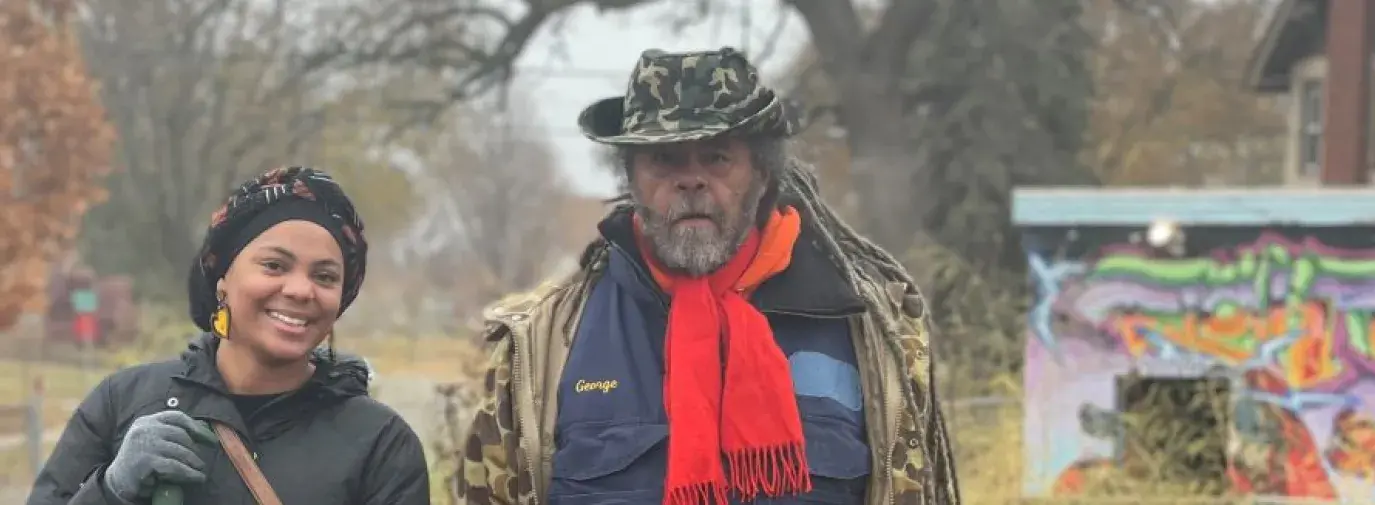 Michael Chaney is wearing a big brown coat and a red scarf, holding a wheelbarrow filled with foliage for compost. He stands next to Jasmira Colon who is smiling at the camera and holding a shovel with an orange handle.