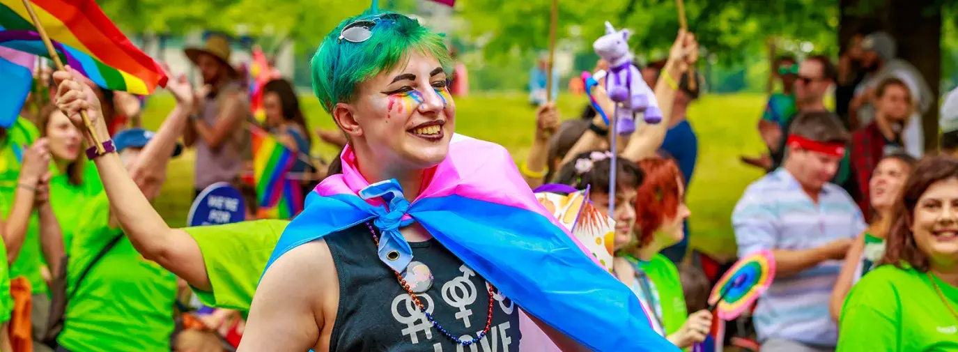 Happy person with rainbow makeup wearing a trans flag as a cape in a parade of rainbow flags.