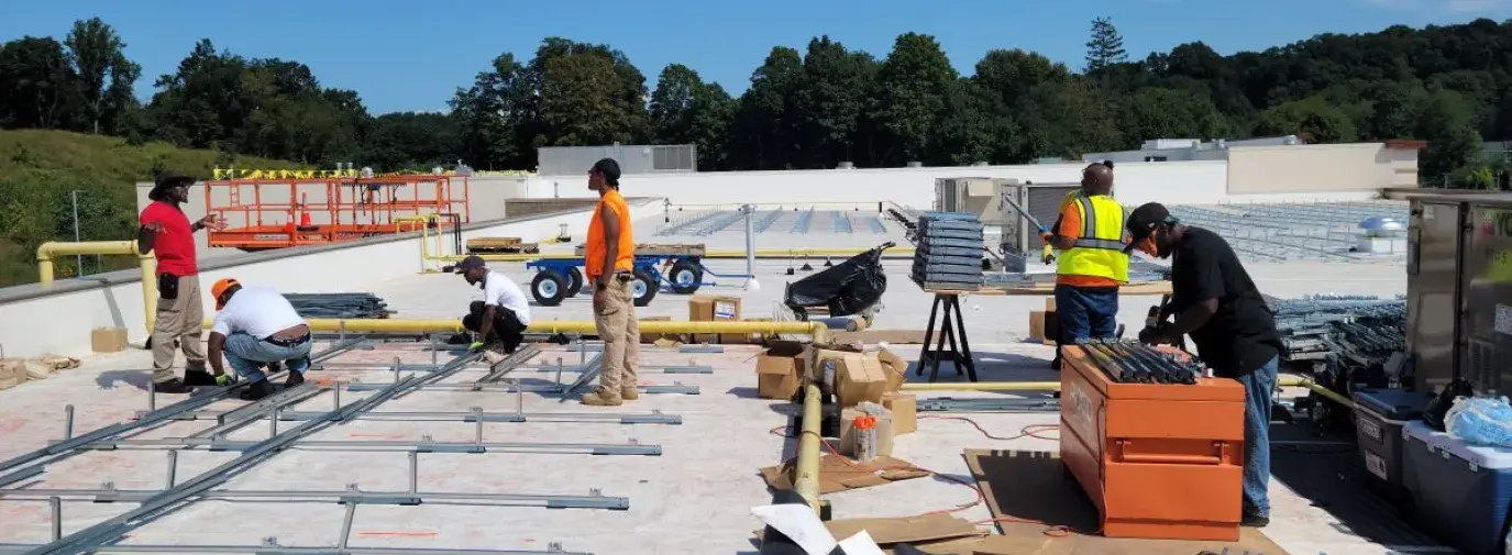 six solar workers standing on a rooftop installing solar panels. Each one is doing something different, whether drilling together metal pieces, supervising, or assembling the pieces together.