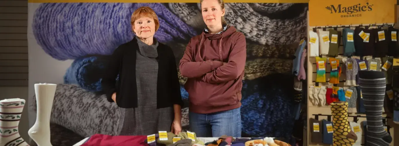 Bená Burda and Sarah Queen stand in front of a display table with socks, yarn, and other fabric items. Behind them is a big poster for Maggie's Organics.