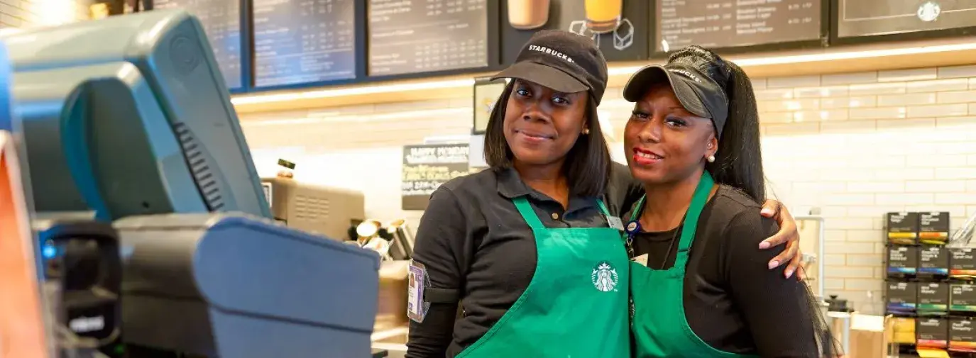 two Black women Starbucks baristas are smiling at the camera. One has her arm wrapped another the other in a side hug. They look prepared and ready for work.