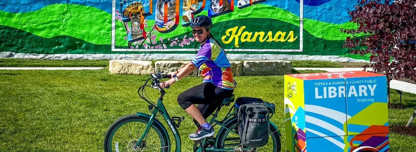 A woman is riding a green bike with a box attached to the back that reads "Topeka & Shawnee County Public Library." She is in front of a mural that says "Greetings from Topeka, Kansas."
