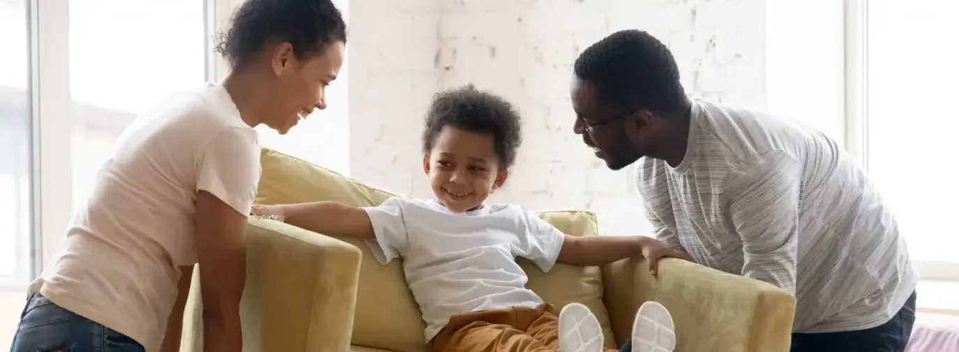 A Black mom and dad pick up an armchair with a 4 year old kid sitting in it. They're all smiling and laughing together.