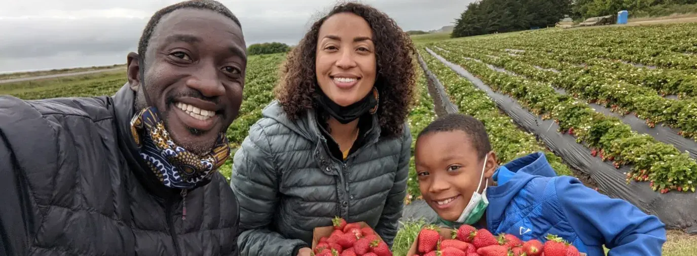 The Nortey family, made up of a man, woman, and son, are smiling at the camera while each holding a box of strawberries. They are standing in front of a strawberry field. 