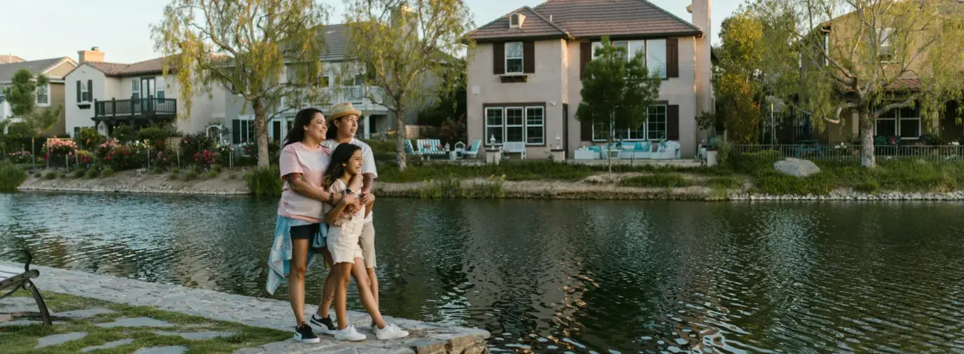 Two women stand with their arms around a young girl, their daughter, at the edge of a small lake with homes and trees in the background. Clean energy for all.