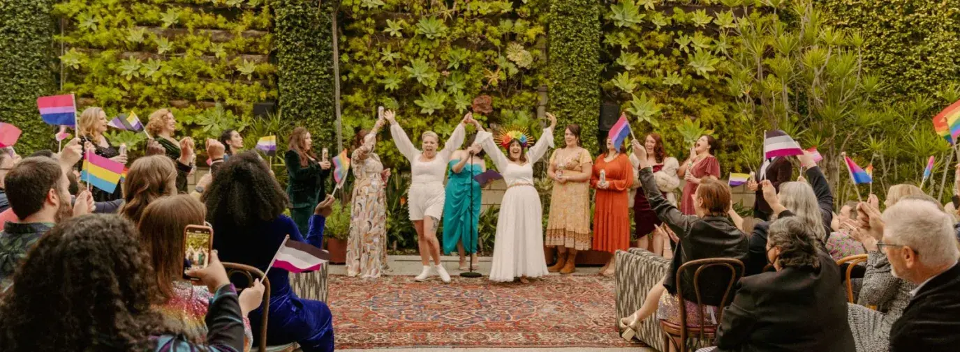 Dana and Anya raise their arms triumphantly during the wedding ceremony. They are surrounded by loved ones waving pride flags and a green, leafy background.