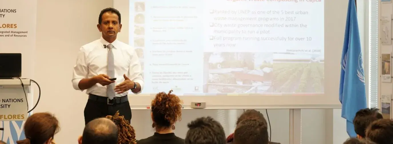 Hiroshan Hettiarachchi is giving a seminar, speaking to a class of diverse people that are all facing him. Behind Hettiarachchi is a powerpoint and to the far right is the UN flag.