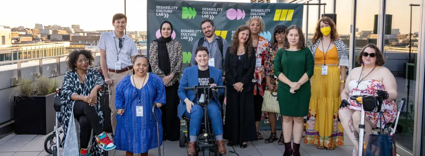 Twelve people of varying diversities and disabilities pose with canes, wheelchairs, and scooters in front of a Disability Culture Lab banner to celebrate the launch of the nonprofit.