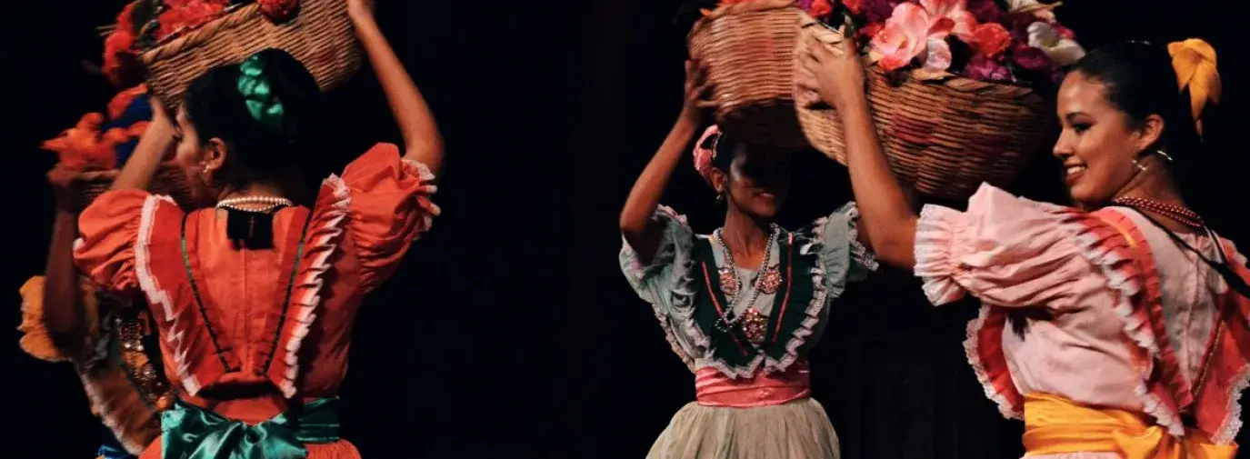 Women in traditional Mexican dresses dancing in a circle while holding baskets above their heads.