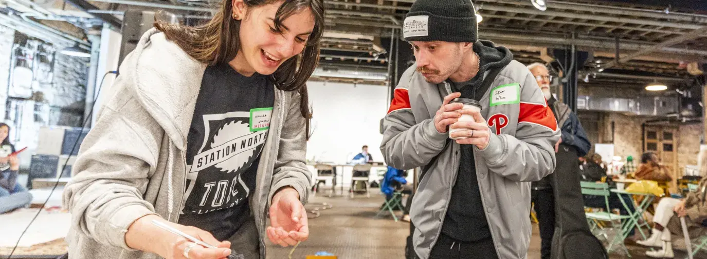 A woman with a staff shirt is explaining how to repair and clean a violin. A man holding a coffee cup is watching her work.