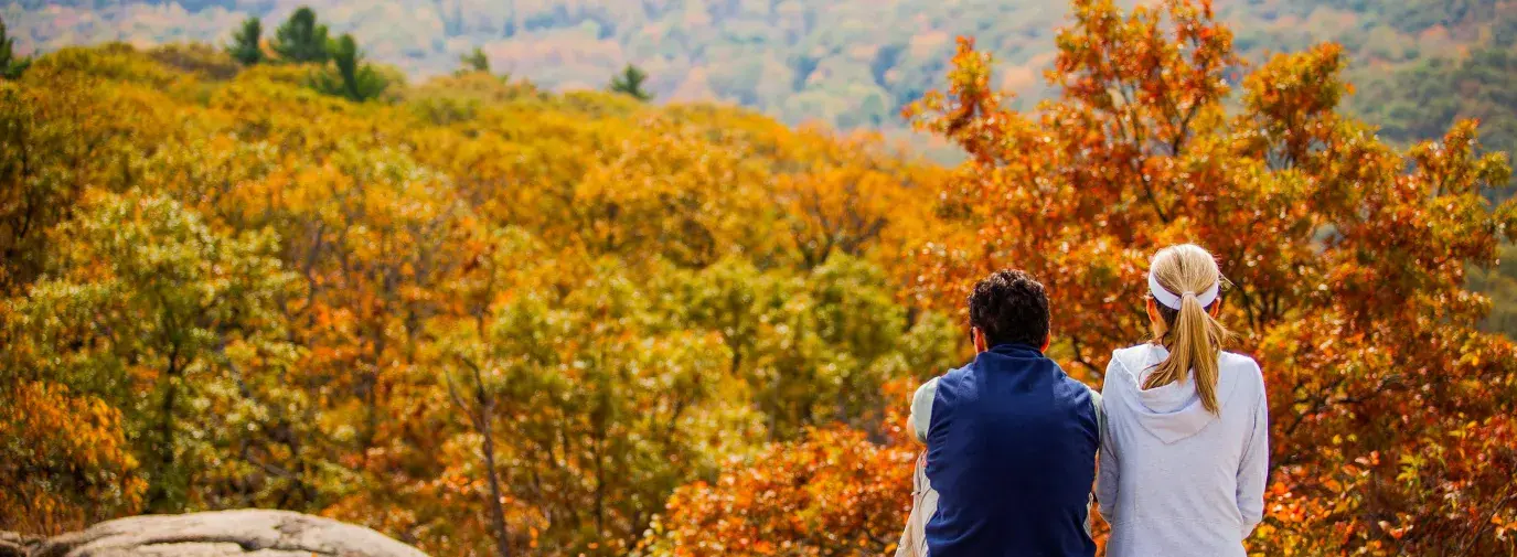 Two people sitting on a rock outcrop overlooking a vibrant autumn landscape of orange and gold trees with misty mountains in the distance