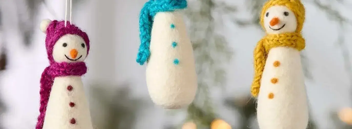 Three handmade felt snowman ornaments with colorful knit scarves in magenta, turquoise, and yellow hanging against evergreen branches.