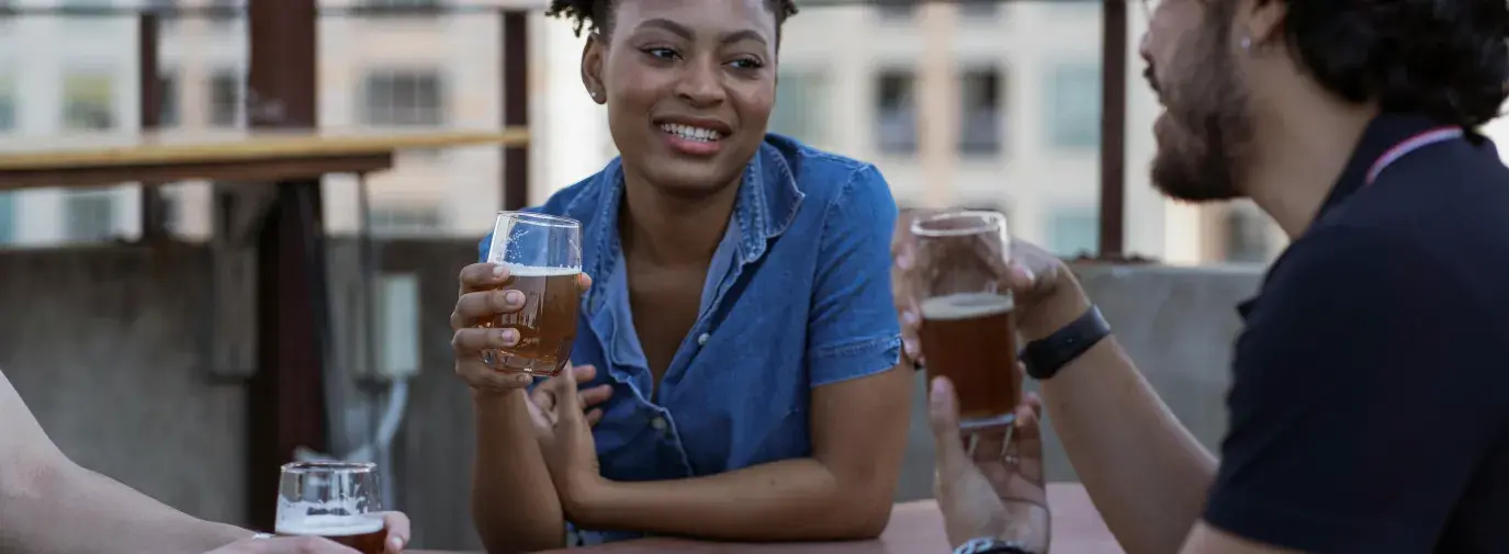 Woman smiling while holding a beer glass at a rooftop bar with two friends, urban buildings visible in the background.