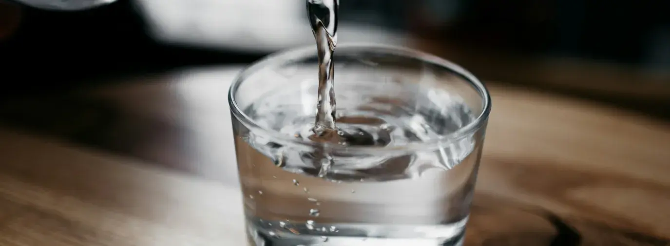 Water pouring from a glass bottle into a glass jar, which is set on a wooden table.