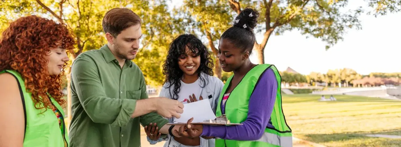a group of four young people wearing construction vests and button down shirts, looking at a clipboard together. They all have curious and engaging expressions. They are standing outside and its sunny and green.
