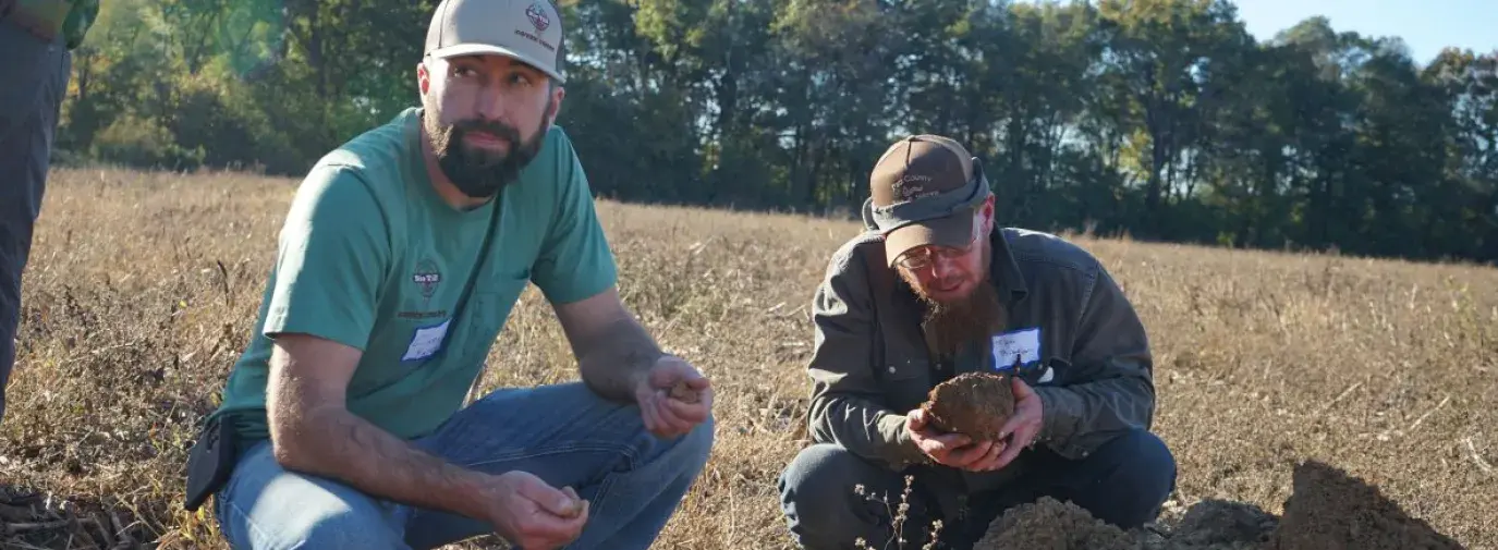 two men squatting in the dirt of a field, holding clumps of soil in their hands and examining them.