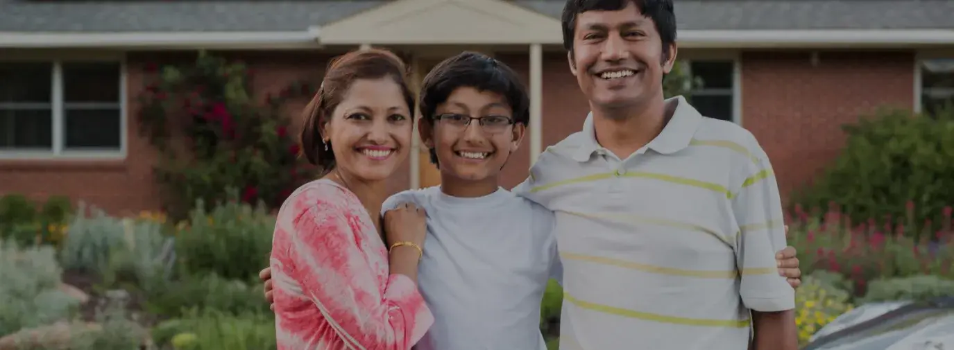 A southeast asian family unit. Mom is wearing a tie dye pink shirt, 9 year old son is wearing glasses and smiling, dad is wearing a yellow striped shirt. They are all hugging each other and smiling at the camera.