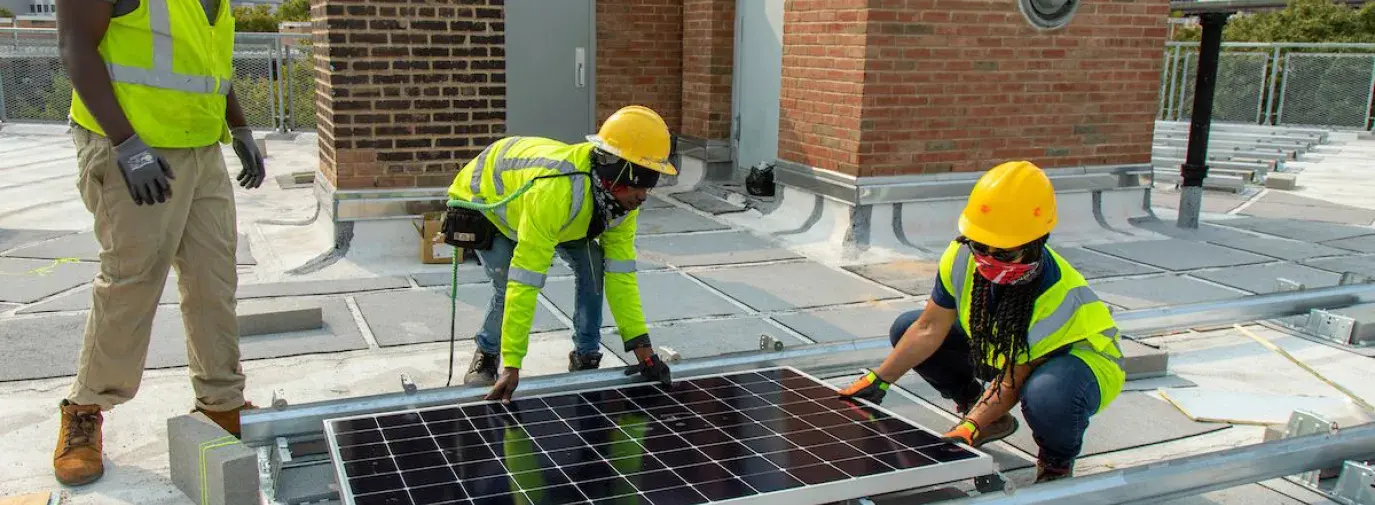 three men in yellow vests wearing hard hats working on installing a solar panel on a roof.