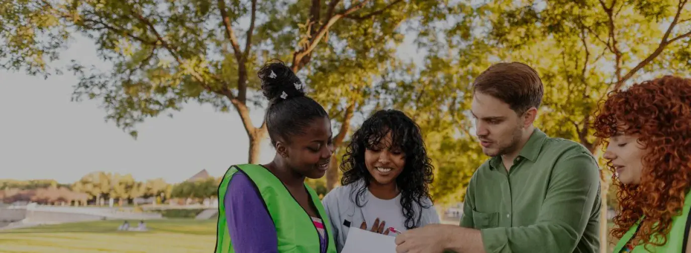 four young adults dressed in neon construction vests discussing a plan on a clipboard.