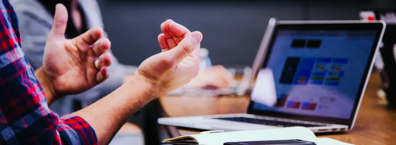 person sitting at a table in front of a laptop, gesturing with their hands, a woman sits in the background, root & leaf specializes in marking consulting