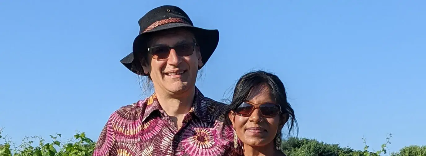 Swati & Mark standing together against a green landscape. Swati is wearing a bright orange dress and they are both smiling.
