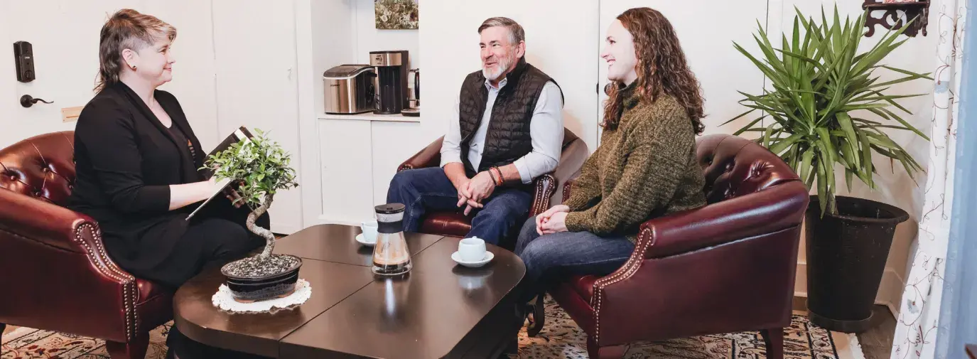 Athens Impact Socially Responsible Investments' founder Michelle Williams sits in a brown leather armchair - on her right are two clients, a white man in a black vest and a white woman with long brown hair in a green sweater, sitting in brown armchairs. Michelle has short hair, wears a black pantsuit, and has a folder in her hands.