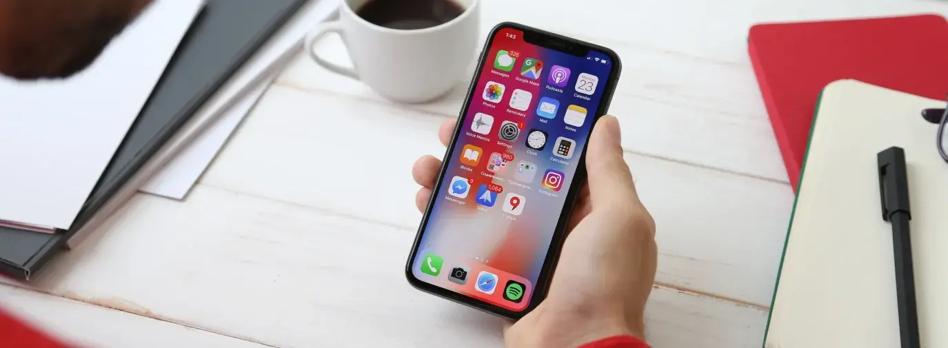 A iPhone, showing the home screen and apps, held in a hand atop a desk with a cup of coffee and a notebook and pen.