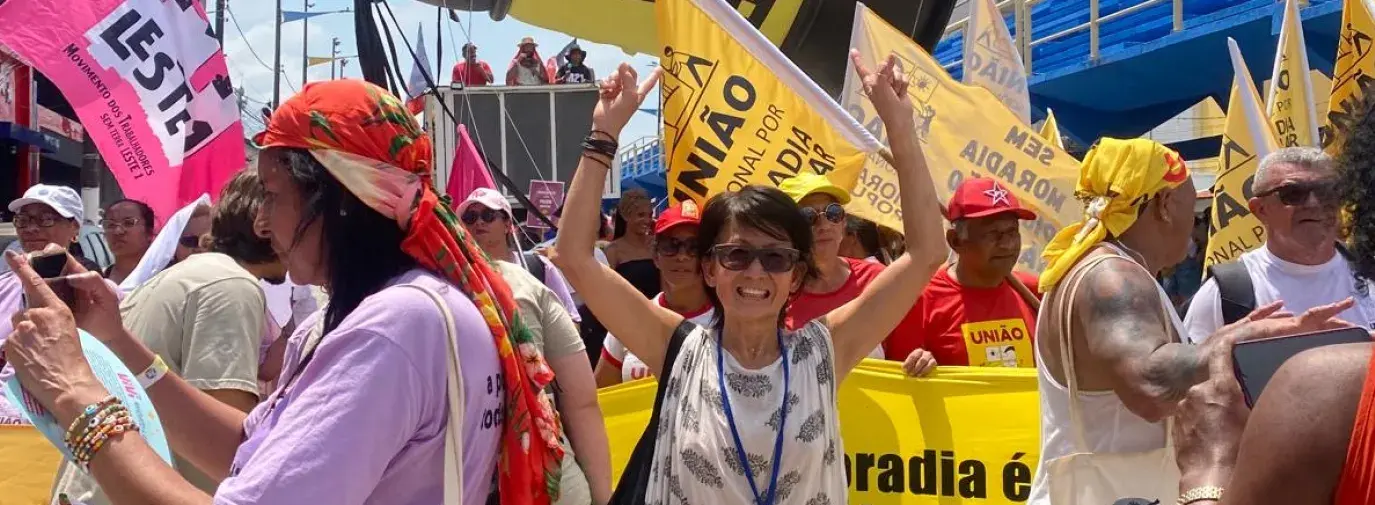 A woman in a patterened white dress stands amidst climate protesters with her arms raised victoriously.