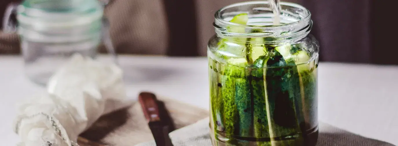 A hand holding a blue pot pouring liquid into a mason jar full of pickling cucumbers.