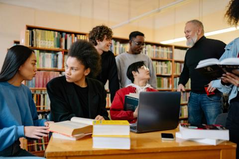 A diverse group of people at the library, talking, with books and a laptop open at the table.