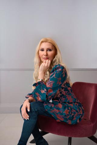 A white woman sits on a burgundy velvet chair against a white wall. The woman has long blonde hair and is wearing dark teal pants with a patterned button up. Women Business Owners.