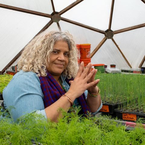 Erika Allen is a Black woman with blonde curly hair. She is smiling coyly at the camera with her hands together while surrounded by seedlings.