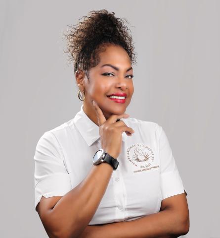 Sylvia M. Walker, a Black woman in a white button shirt. Her left arm is across her stomach, her right arm is bent at the elbow, her chin in her right hand which sports a watch. Women Business Owners.