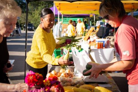 Three people are shopping at a farmers market stand, picking produce. A Black woman in a yellow sweater picks an ear of corn. A middle-aged white woman picks cucumber to put in her white plastic bag. An elderly woman is looking at flowers.
