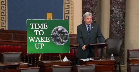 Sen. Sheldon Whitehouse (D-RI) giving a speech at a podium with a green sign that says Time To Wake Up next to him. 