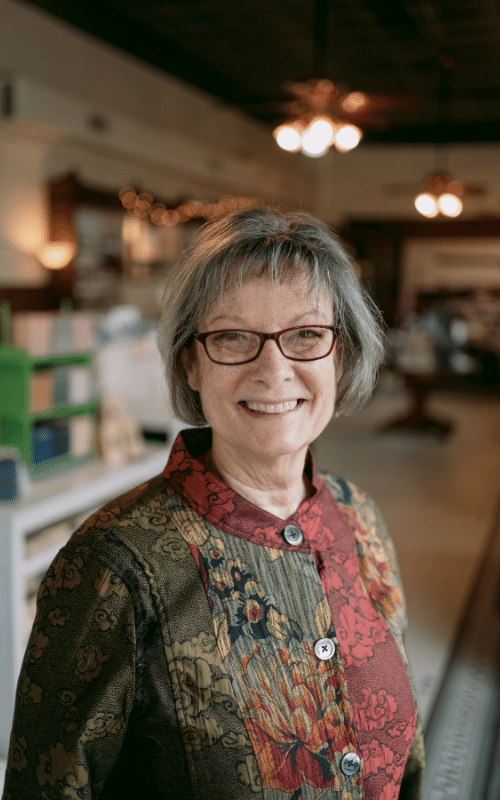 Shelley Green, an older white woman with a greying bob and brown, rectangle glasses, stands center from the chest up in her organic tea store.