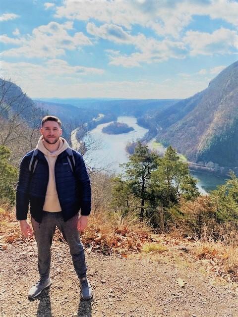 A man in sneakers, grey pants, a white hoodie, and a navy puffer jacket stands outside with a massive river behind and below him. Gen Z finances.