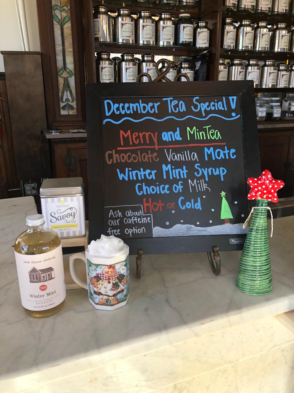 A hand-written sign on a counter with a wall of tea behind it. The sign reads: "December Tea Special! Merry and MinTea: Chocolate vanilla mate winter mint syrup choice of milk hot or cold." Organic tea.