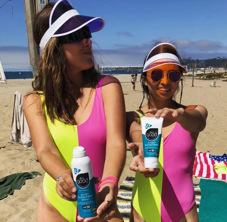 Two women in neon pink and yellow one piece bathing suits, wearing visors, hold out mineral sunscreen. Sustainable summer.