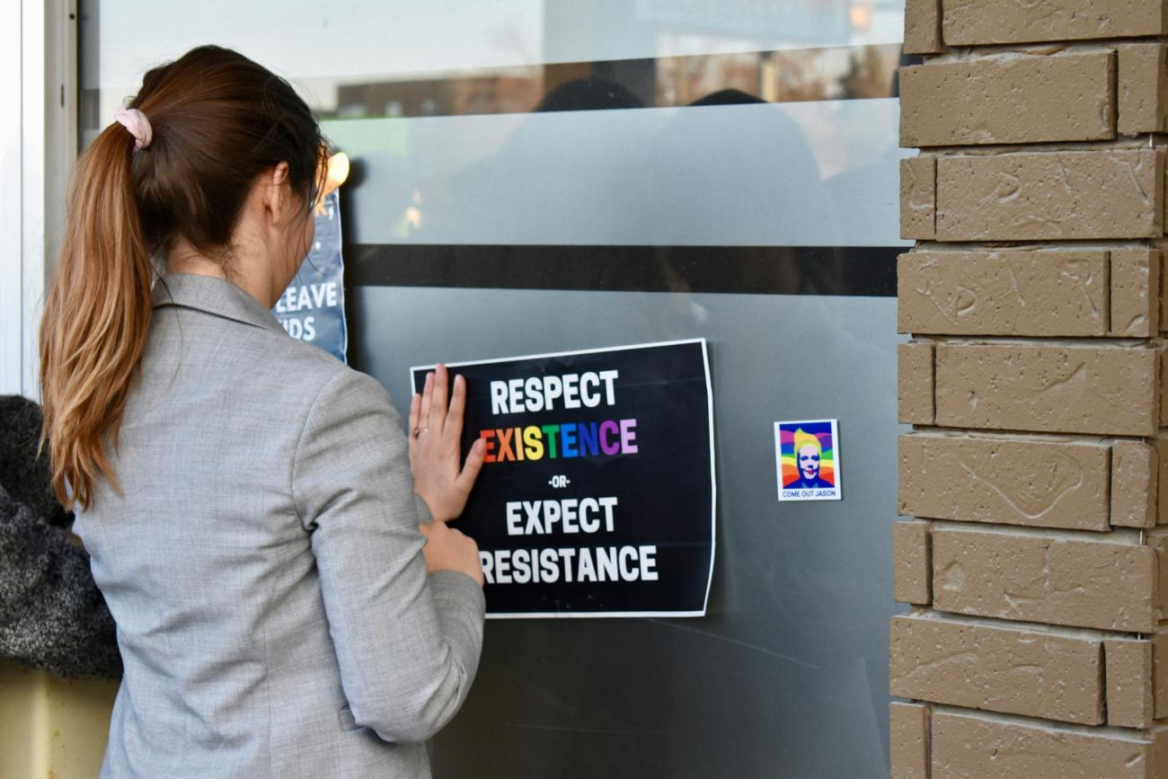 A person shot from behind, wearing a grey blazer, brunette hair up in a ponytail. They put a sign up on a window of a building that reads: "Respect existence or expect resistance."

The word "existence" is in rainbow letters.
