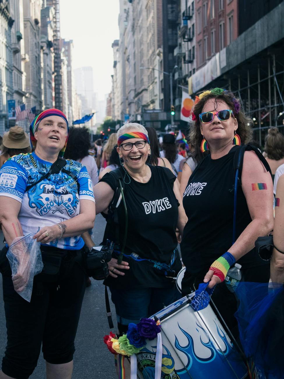 Three white people stand together at Pride, wearing various rainbow accessories like headbands and gloves. Two of them wear shirts that read "Dyke."